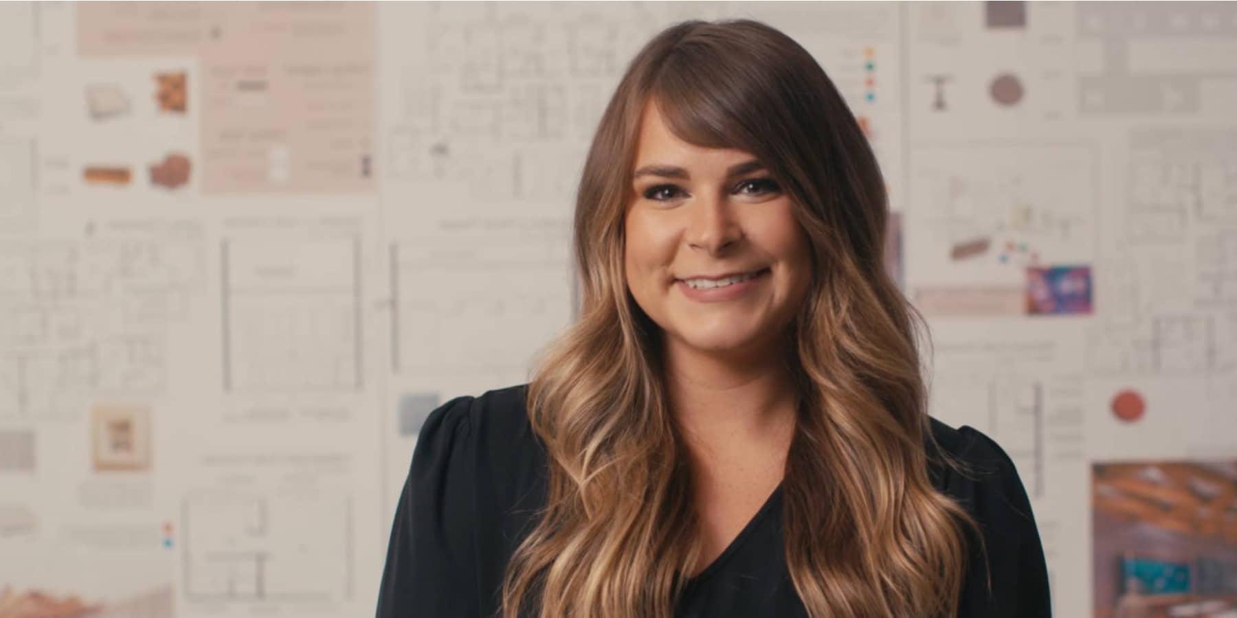 A woman with wavy brown hair poses with interior design sketches and designs behind her. She wears a black dress shirt.