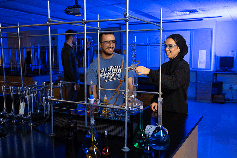 Three students of different ethnicities stand in a blue-lit lab. On the left is a female student wearing a black outfit and black hijab. To her right is a male student with short brown hair. He wears a blue Indiana State T-shirt. Behind them is a male student in a black jacket. The students are wearing safety glasses, and the female student is pointing to a long, tall test tube.  