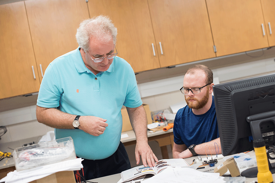 Two men are in a classroom. On the left is a man with short brown hair, with a matching beard and mustache, and glasses. He sits at a table, wearing a dark blue T-shirt. On the right is a man with short white hair and glasses. He is standing and wearing a light green polo shirt. On the table in front of them is an open textbook and other papers, with a desktop computer to the left. Brown cabinets are behind them.