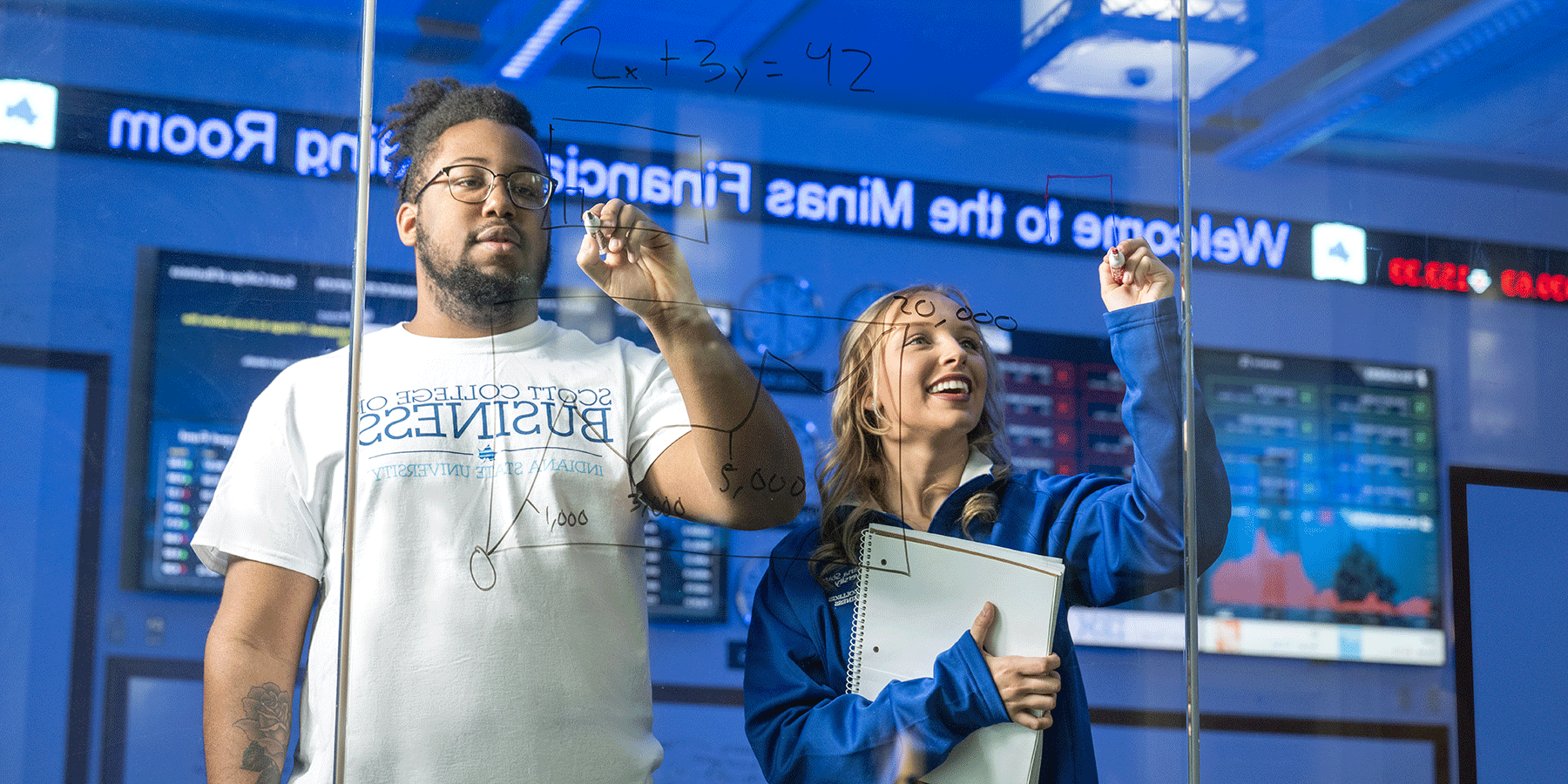 Two students write on a clear dry erase board with news ticker in background