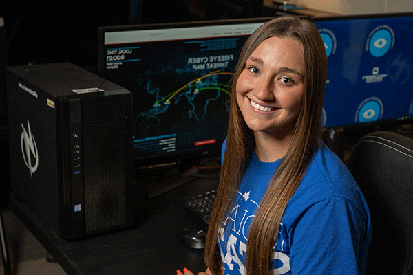 A white female student with long brown hear wearing an Indiana State shirt looks back at the camera while sitting in a dark room with a computer with a threat analysis website on the screen. 