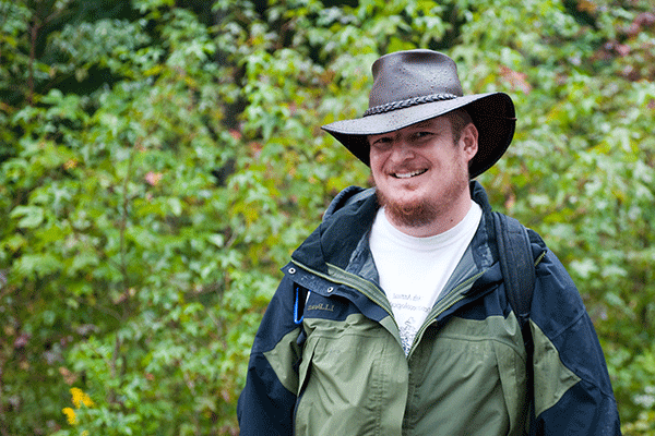 Portrait of a white male professor with mustache and beard wearing a hat and light jacket with green foliage in the background.