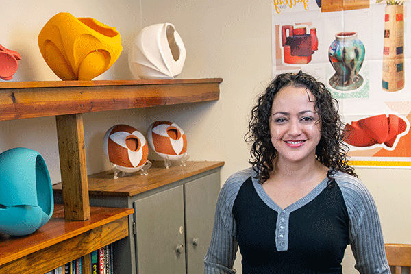 A smiling Latina woman with curly black hair and wearing a black-and-grey, long-sleeved jersey t-shirt with buttons stands in a room with colorful pottery pieces on shelves beside her. Visible behind her is a poster that illustrates examples of other pottery designs.