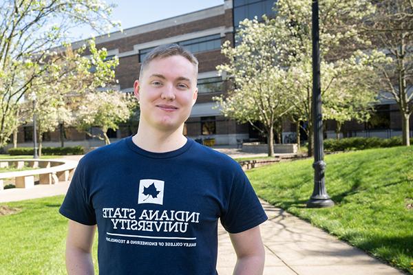 A white, male student smiles as he stands at some distance in front of a multi-story brick building, the John T. Myers Technology Building. The student has short blond hair and is wearing a navy blue t-shirt that says “Bailey College of Engineering & Technology” on it. It also has the blue shape of a Sycamore leaf logo in a white box. In the background, green grass and trees with white flowers can be seen.