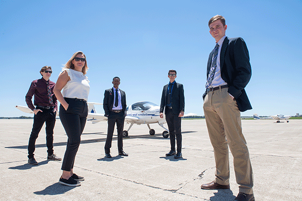 Four men and one woman stand on an open airport tarmac. A white airplane with blue lettering that says Indiana State University is visible behind them. The sky is blue and the day is bright and sunny. Another white plane is visible far in the distance on the left of the image.