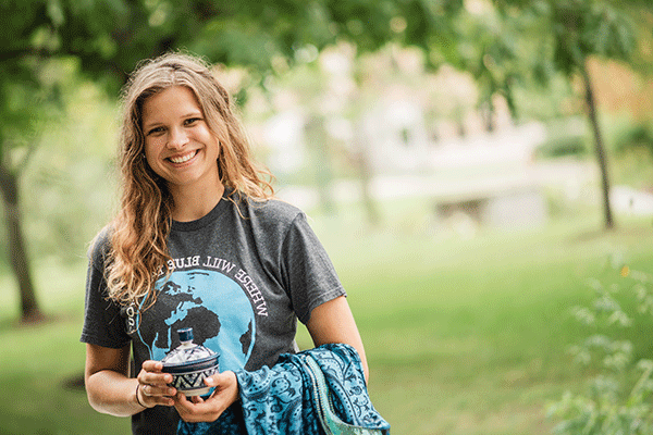 A white female student with long curly light brown hair poses outside. She wears a grey T-shirt with a globe showing North and South America in blue colors. “Where Will Blue” in white lettering is visible above the globe. She holds a blue jacket in her right hand and holds a blue-and-white ceramic cup with a lid in her left hand. Green trees are visible in the background, blurred in the photo. 