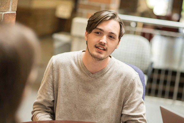 A smiling white male student with sparse facial hair and dark brown hair that falls left across his forehead is seated and talking to someone who is only visible from the back and is blurred. The man wears a beige sweatshirt. Behind him, out of focus, can be seen other tables, chairs, a brick pillar, a metal railing, and other elements of the commons area in the student union.