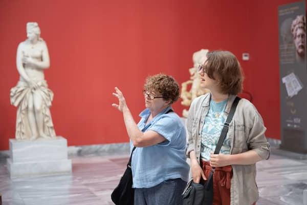 Two individuals are standing inside an art gallery with a red wall in the background.