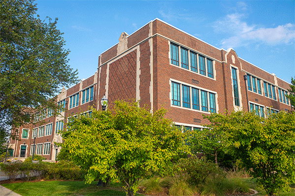 Exterior image of a large, multi-story brick building that houses the Bayh College of Education with green trees obscuring the lower half of the image with a blue sky.  