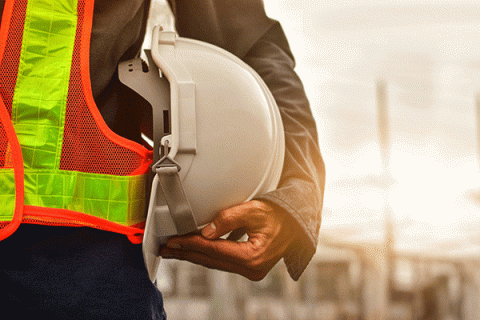 A person holding a white construction hat while wearing an orange-and-yellow safety vest over a grey long-sleeved shirt, and dark pants. A grey-and-white background with indistinct structures is blurred behind the individual.