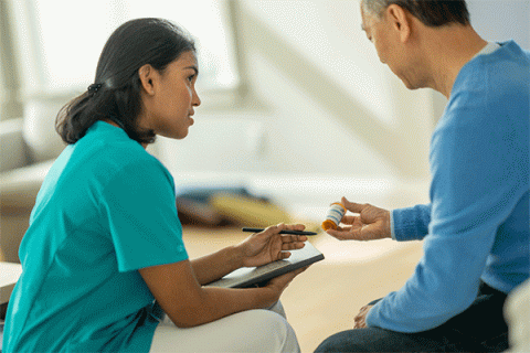 A senior gentleman of Asian descent, sits in a living room in his home with his nurse practitioner, during a home care visit. He wears a blue long-sleeved shirt and dark blue jeans. He holds a bottle of prescription pills. The practitioner is a tan-skinned woman with dark black hair pulled back behind her shoulders. She wears teal scrubs and khaki pants.  